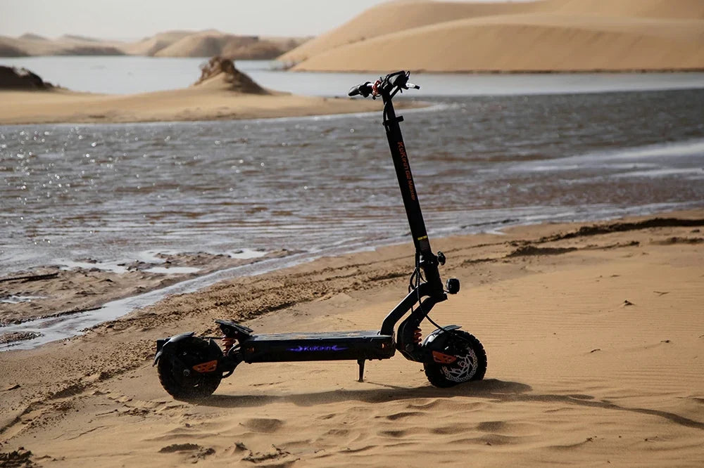 Atmospheric static product shot of black-orange Kukirin G2 Master electric scooter propped on desert sand beside a lakeside with rolling sand dunes in the backdrop.