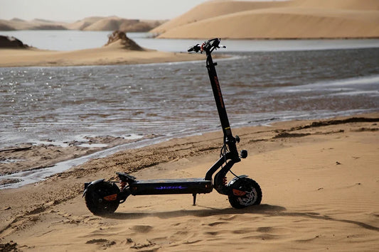 Atmospheric static product shot of black-orange Kukirin G2 Master electric scooter propped on desert sand beside a lakeside with rolling sand dunes in the backdrop.