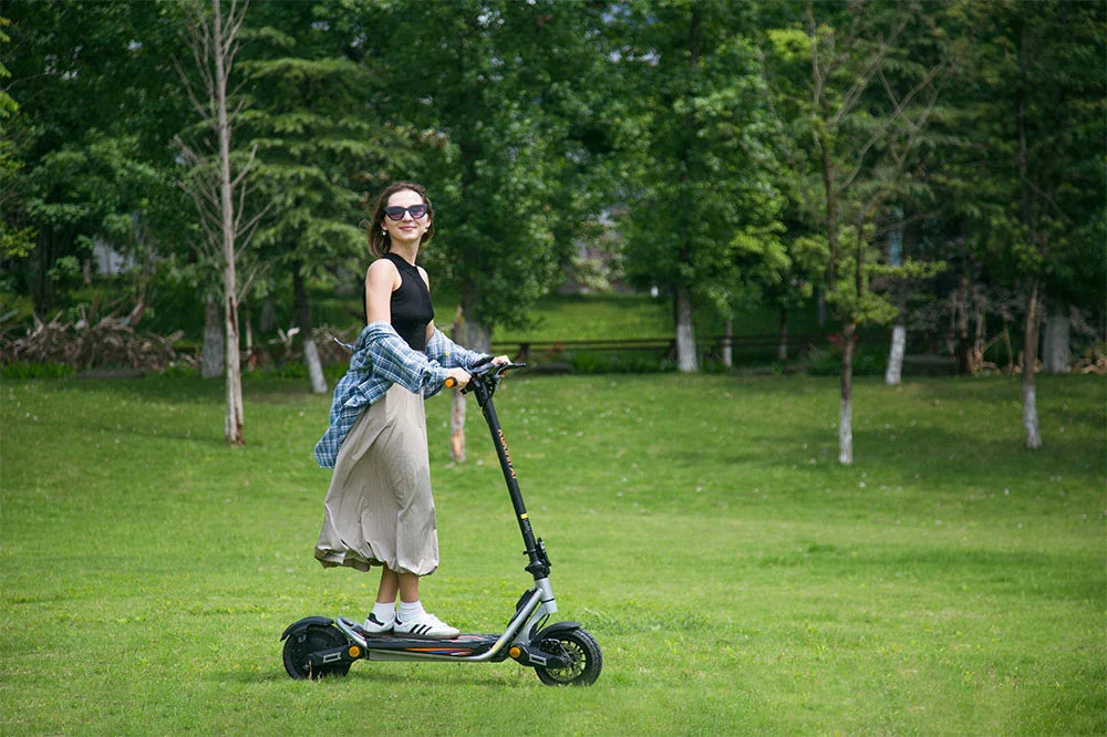 Silver-black Kukirin lightweight folding e-scooter, female rider cruising lush grass surrounded by dense green trees for casual outdoor leisure