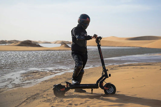 Desert oasis action shot of fully protected rider standing on black-orange Kukirin G2 Master foldable electric scooter on golden lakeside sand dunes.