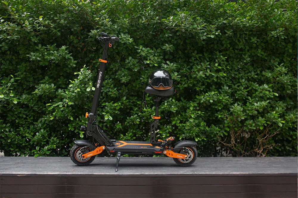 Static product showcase of black and orange Kukirin G2 Max foldable electric scooter with a helmet resting on the seat, placed on dark stone ledge against dense lush green hedge backdrop.