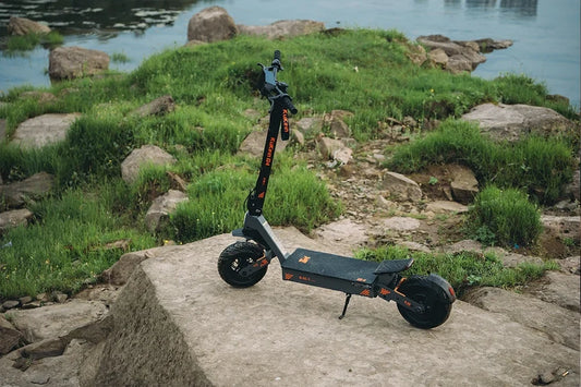 Static outdoor shot of Kukirin G4 off-road electric scooter parked on large stone beside river, green grass and rocky shore background.