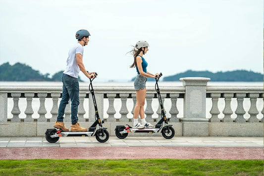 Man and woman riding Kukirin M4 Max electric scooters side‑by‑side on seaside promenade
