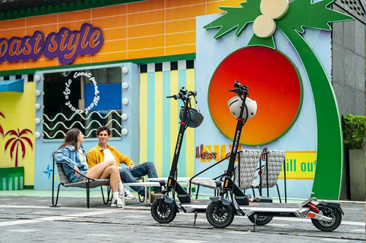 Two Kukirin M4 Max electric scooters parked in vibrant colourful urban street, young couple relaxing on background seating