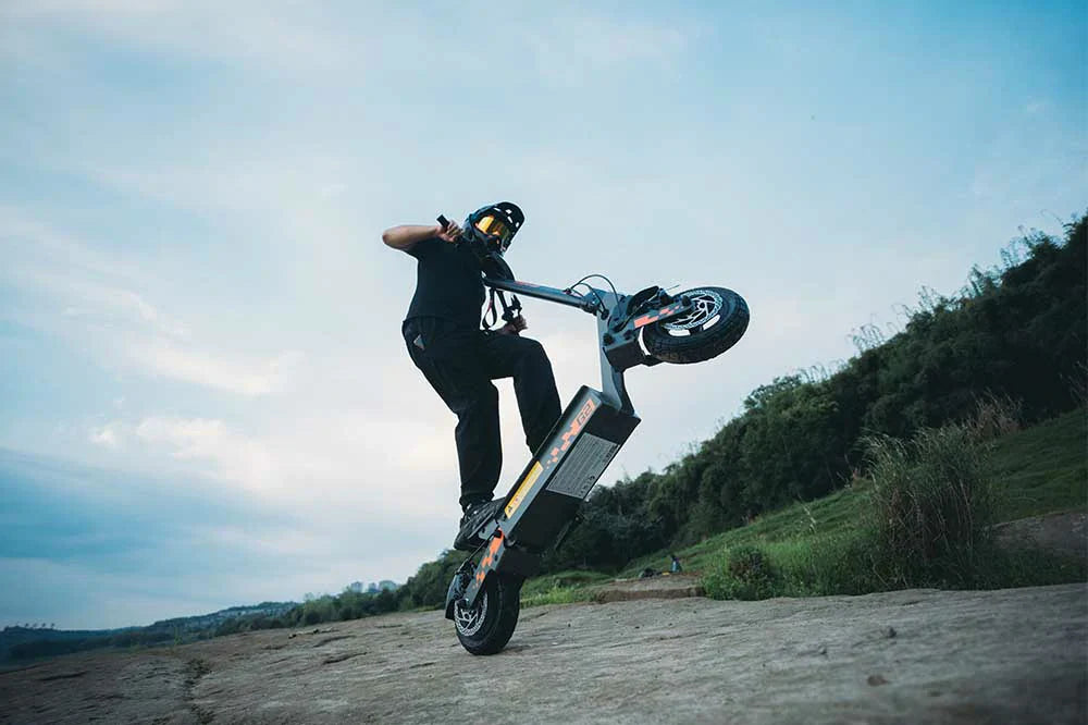 Black-orange Kukirin G2 rugged dual-wheel e-scooter, fully geared rider lifting front wheel on rough stone ground surrounded by lush green hills and soft cloudy sky