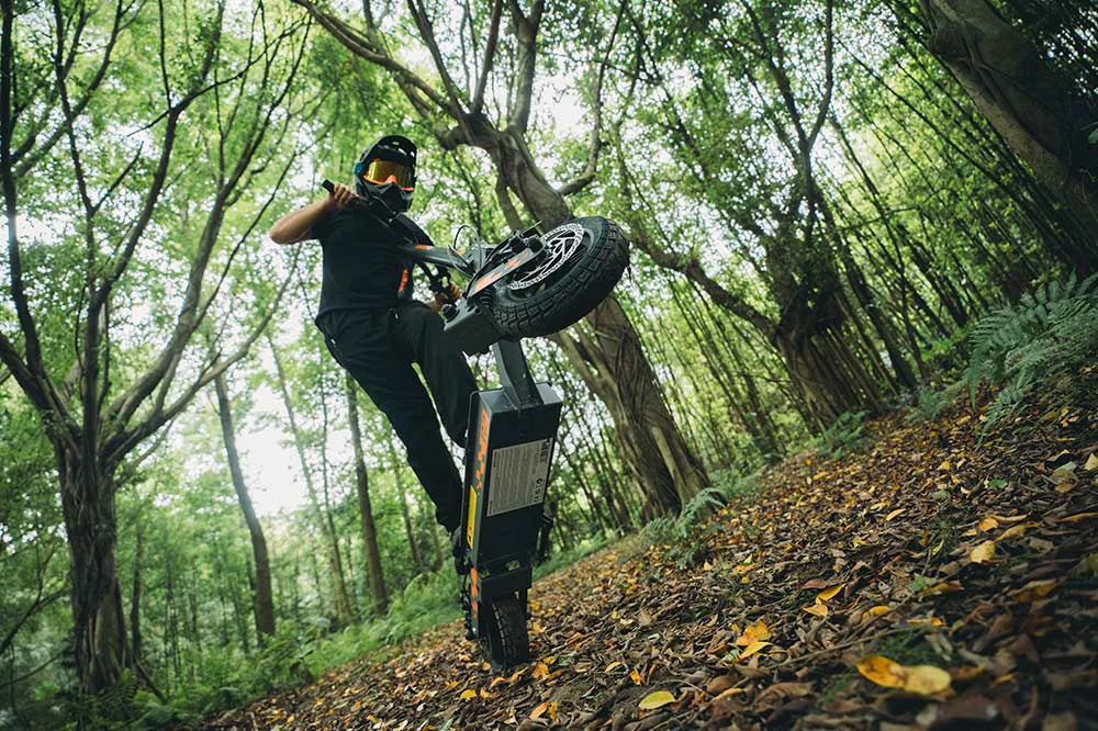 Rider in protective gear performing wheelie on Kukirin G2 black-orange electric scooter on leaf-covered forest trail surrounded by lush green trees