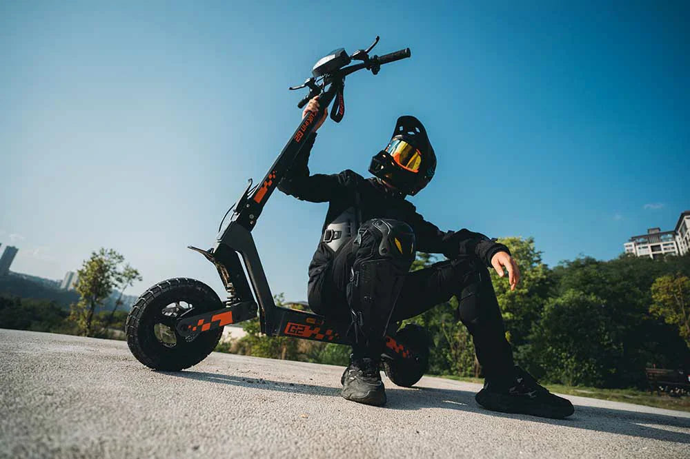 Rider in full black protective gear posing with Kukirin G2 VMP black-orange off-road electric scooter on concrete platform under a clear blue sky, with greenery and residential buildings in the background