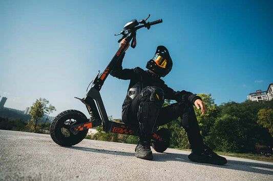 Rider in full black protective gear posing with Kukirin G2 VMP black-orange off-road electric scooter on concrete platform under a clear blue sky, with greenery and residential buildings in the background