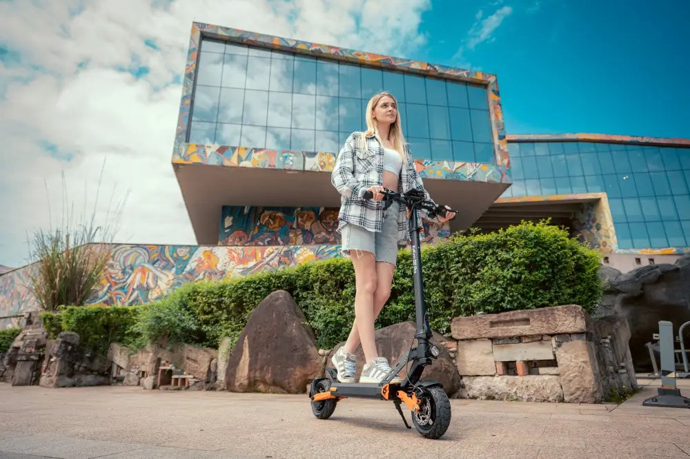 Urban lifestyle shot of blonde woman riding black orange Kukirin G2 Pro foldable electric scooter outside modern graffiti art museum building.