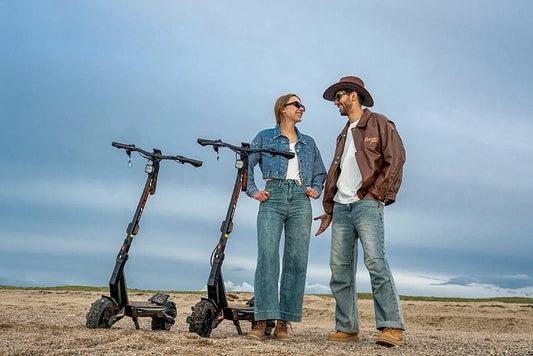 Coastal beach lifestyle shot of a smiling young couple standing beside two black off-road Kukirin T3 electric scooters on gravel sand under a cloudy pale blue sky.